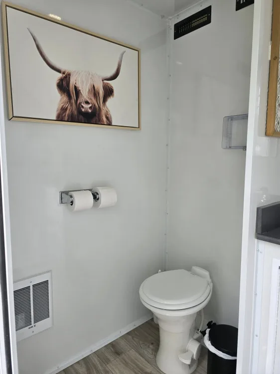 Private stall inside a Restroom Ranch trailer with a porcelain toilet, wood-look floor, and framed Highland-cow artwork